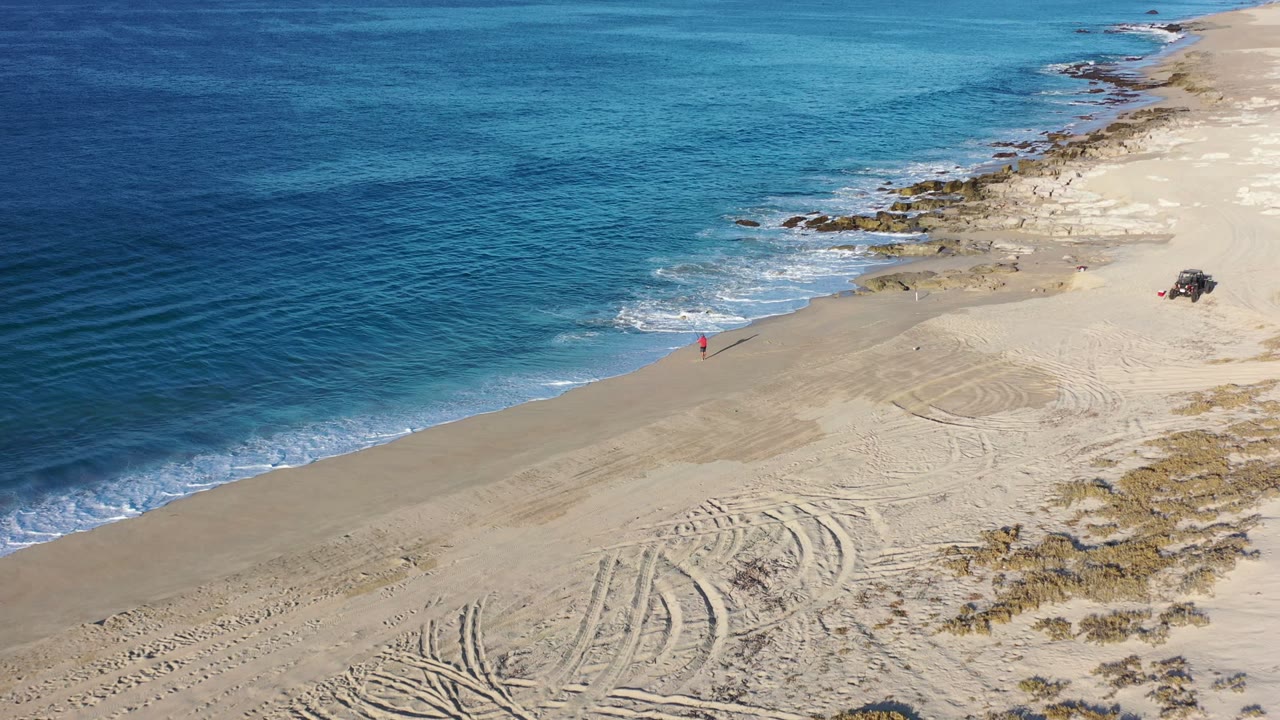 Aerial frame from Los Cabos at 0:01, showing turquoise water pushing onto the beach, sandy tire marks in the lower frame, low shoreline rocks, and a small red figure farther up the sand.