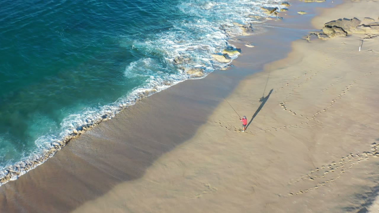 Aerial view of a Los Cabos beach with turquoise water meeting tan sand, small shoreline rocks, footprints, and a tiny distant figure in pink near a long shadow.