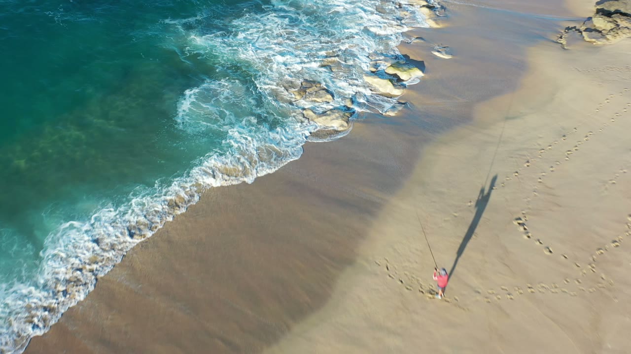 Aerial Los Cabos shoreline frame with foamy surf curling across wet sand, scattered rocks near the waterline, footprints, and a small distant figure low in the frame.