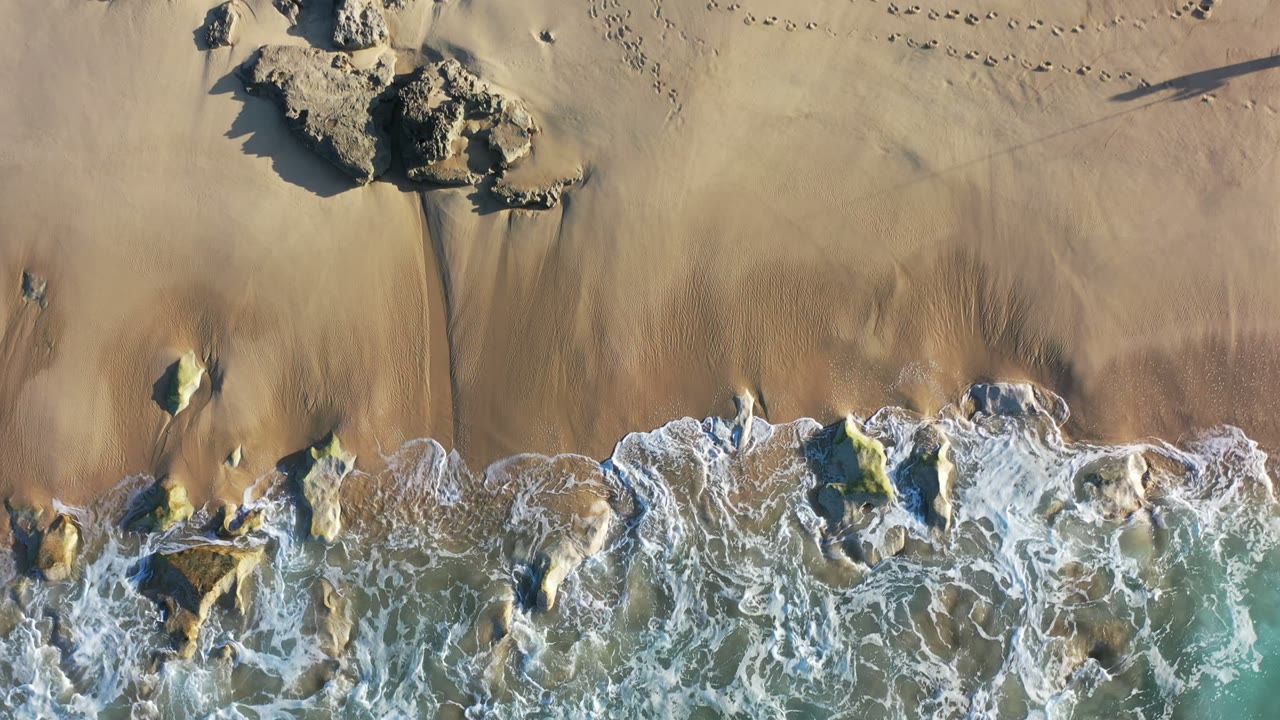 Overhead beach view with rocks, gold sand, and surf foam.