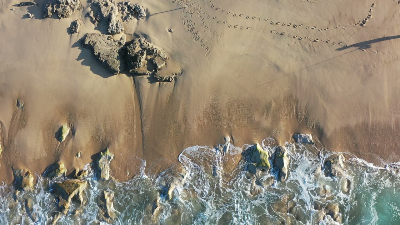 Los Cabos overhead beach view with foamy water wrapping around rocks, broad gold sand above the surf, footprint trails, and no visible people.