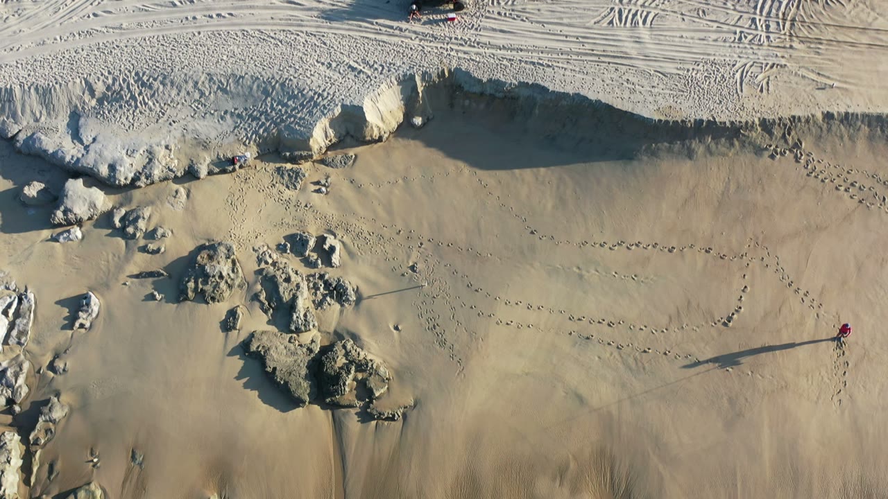 Overhead sand tracks, rocks, and a lone walker near eroded sand banks.
