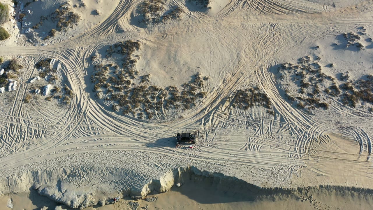 Overhead Los Cabos dune frame with pale sand, curving tire tracks, scrub patches, rocks, a small dark off-road vehicle near the lower left, and no visible people.