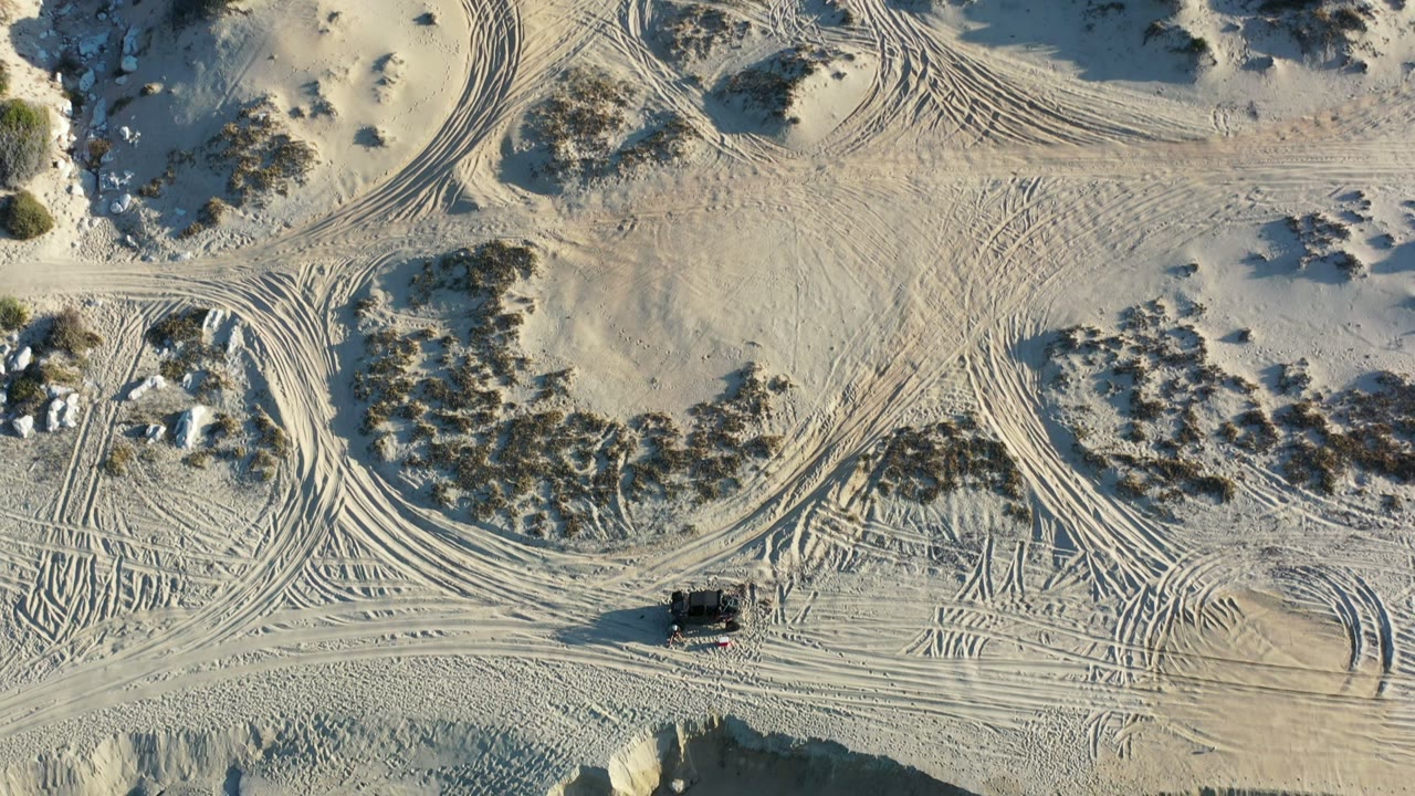 High overhead Los Cabos view of pale dune terrain where multiple tire tracks cross around scrub-covered mounds and rocks, with no visible people.