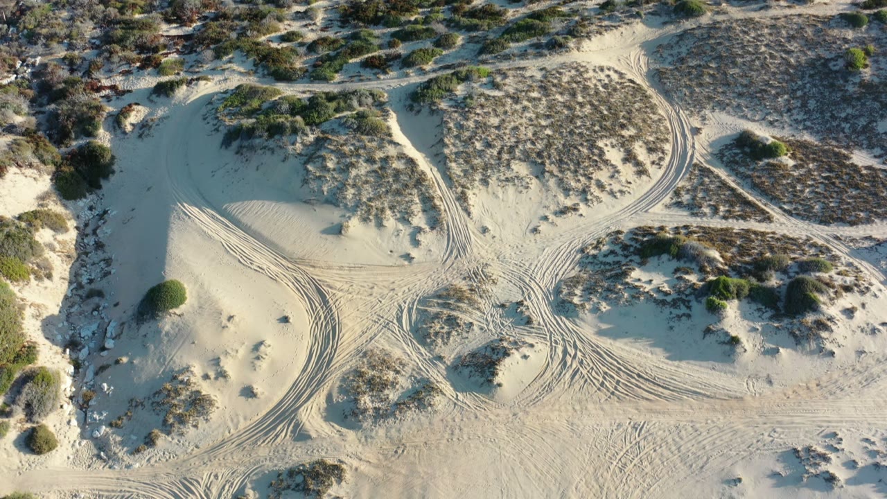 Los Cabos aerial frame with a broad pale sand patch, low shrubs, intersecting tire marks, and no visible people near the center of the terrain.