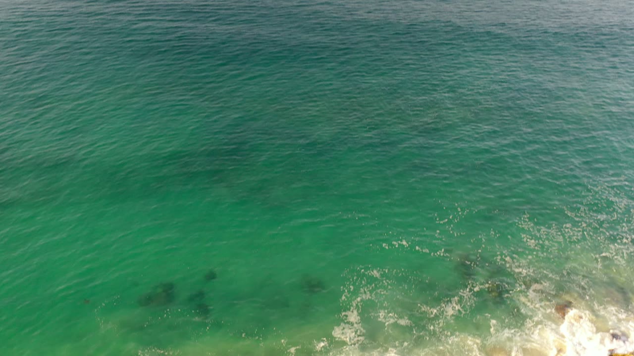 Overhead view in Los Cabos, Mexico, with turquoise water, a faint dark submerged shape near the lower-left area, breaking white surf on rocks, and no visible people.