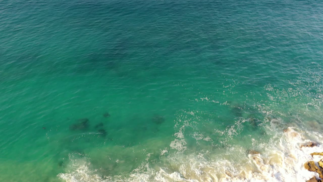 Aerial Los Cabos shoreline frame with green-blue water, darker patches beneath the surface, foamy waves pushing toward tan rocks at the lower right, and no visible people.