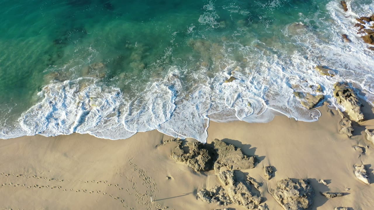 Overhead surf washing onto a sandy beach with rocks and footprints.