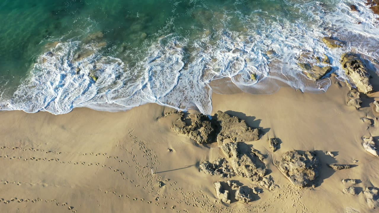 Aerial Los Cabos beach frame with foamy water spread farther over the upper sand, rock outcrops near center, footprint trails below, and no visible people.