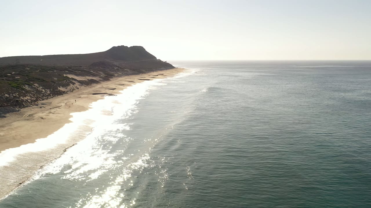 Aerial view of a long Los Cabos beach with white surf, golden sand, dark rocky hills, and no visible people.