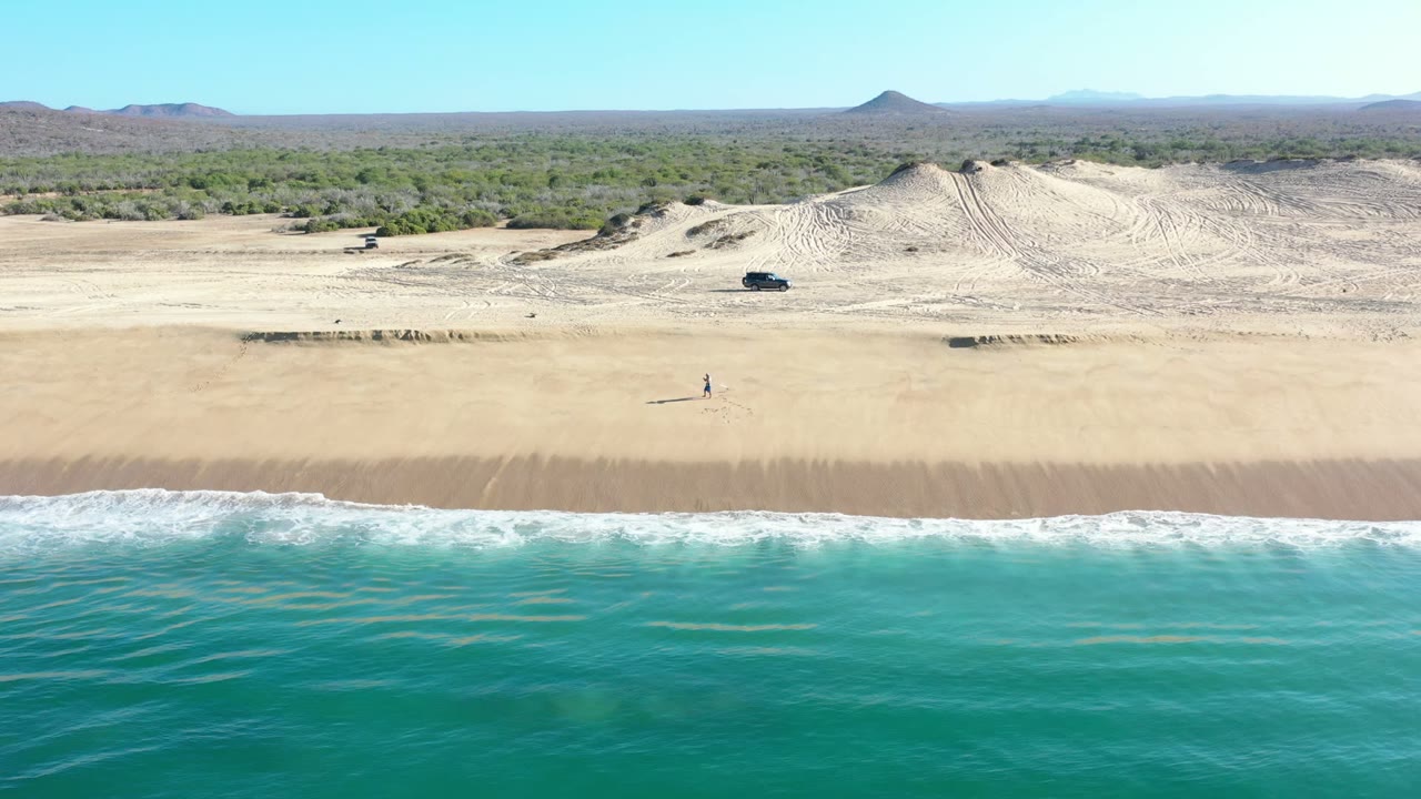 Aerial view of turquoise ocean, sandy beach, dunes, scrubland, and one dark vehicle.