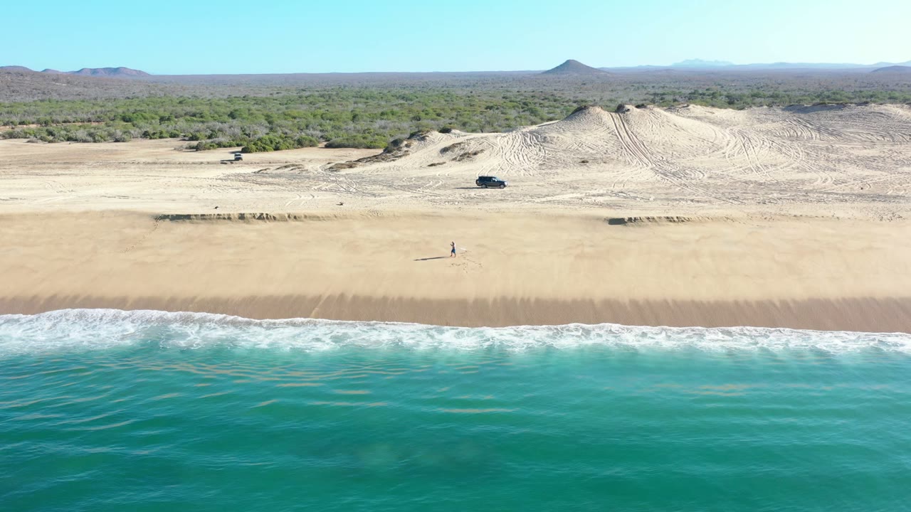 Los Cabos aerial frame showing blue-green ocean across the lower edge, a broad sand strip, low dune mounds, scrubland beyond, and a dark vehicle parked or moving near the dune line.