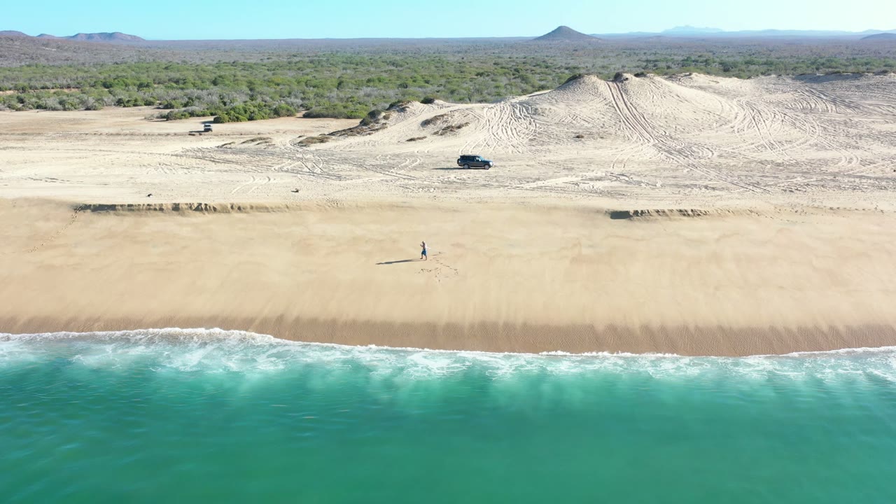 Aerial view of surf, wide sand beach, dunes, scrubland, a dark SUV, and a distant person.