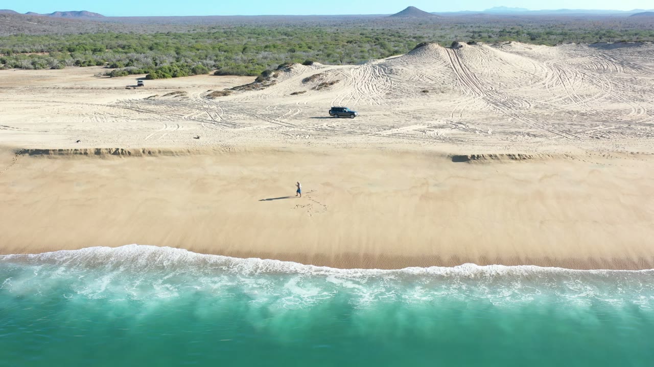 Aerial view of the Los Cabos shoreline with breaking waves along the lower edge, a lone distant person near mid-beach, tracked dunes, scrub, hills, and a dark SUV inland.