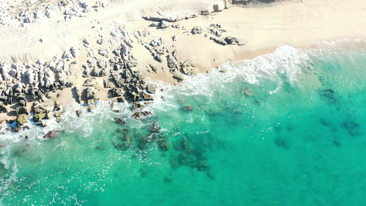 Overhead Los Cabos shoreline frame with turquoise surf, exposed rocks, pale beach, sandy dunes, scrub patches, and no visible people.