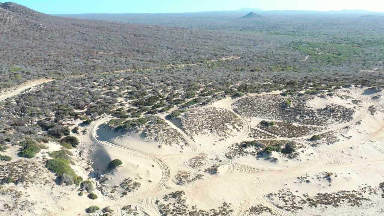 Wide aerial frame over Los Cabos, Mexico desert terrain with pale sand, scattered shrubs, low hills, distant mountain ridges, bright sky, and no visible people.