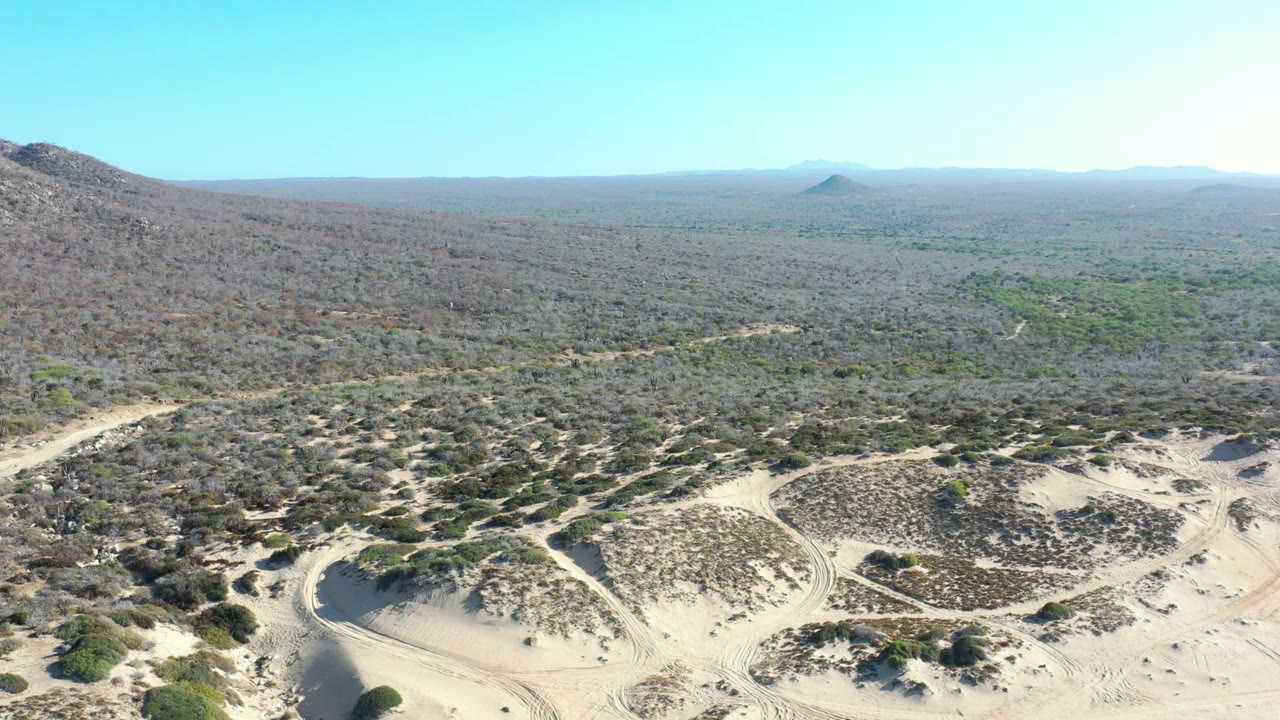 Aerial view in Los Cabos, Mexico with sand flats and scrub in the foreground, a hillside on the left, a distant conical mountain near the horizon, and no visible people.