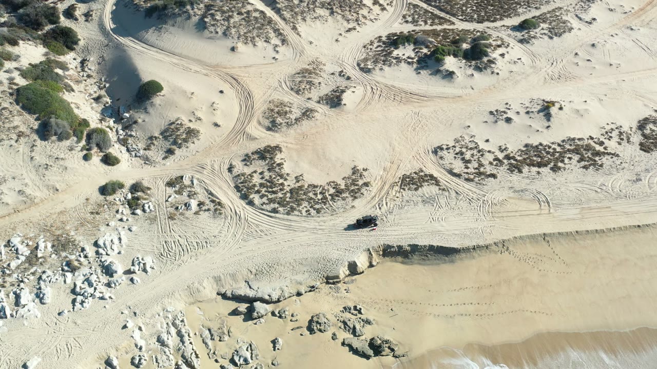 Aerial Los Cabos shoreline frame with looping tire tracks across the dune sand, white rock outcrops near the beach, green water below, and no visible people.
