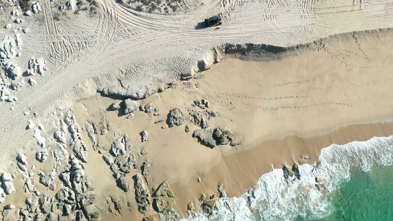 Overhead frame from Los Cabos, Mexico showing pale sand, scattered rock outcrops, vehicle tracks near the top, and white surf entering from the lower edge with no visible people.