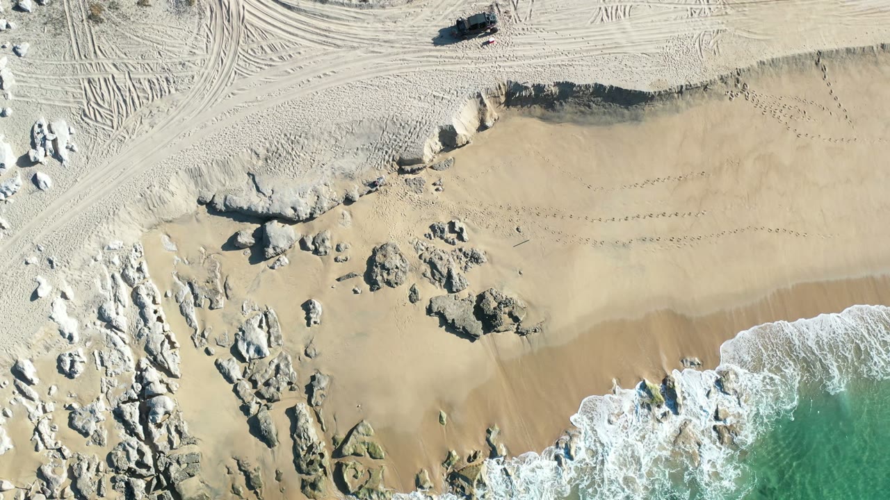 Top-down Los Cabos shoreline frame with surf spreading farther across the lower beach, rock formations along the left and center, tire tracks above the sand, and no visible people.
