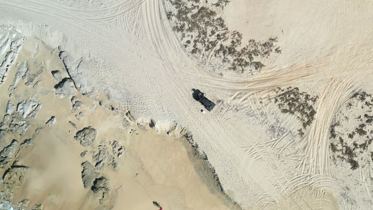 Overhead frame from Los Cabos showing pale sand, rocky shoreline, tire tracks, scrub patches, and a dark vehicle near the beach edge with no recognizable people.