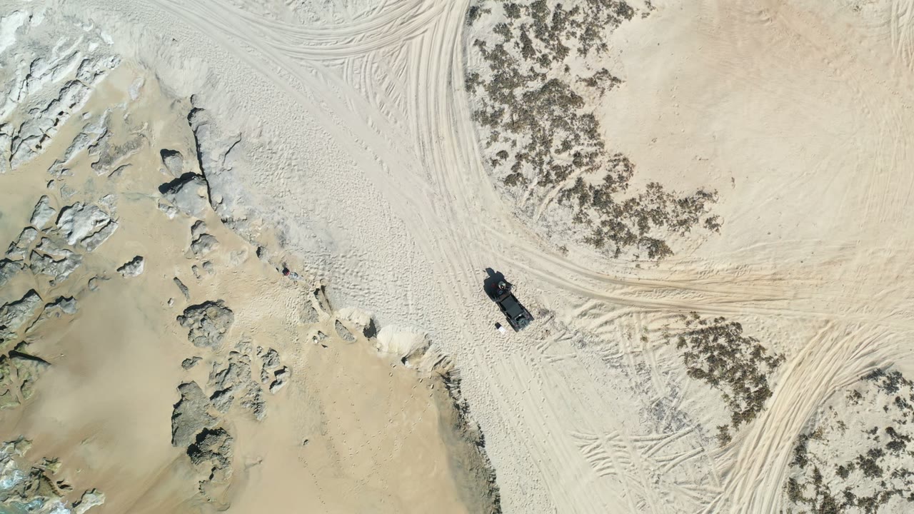 Los Cabos overhead beach view with the camera farther across pale sand, clustered rocks, tire-track patterns, scrub, and a dark vehicle near the upper left of the frame.