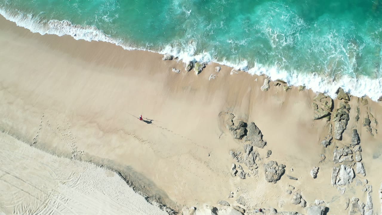 Overhead beach view with surf, rocks, and a small person walking on sand.