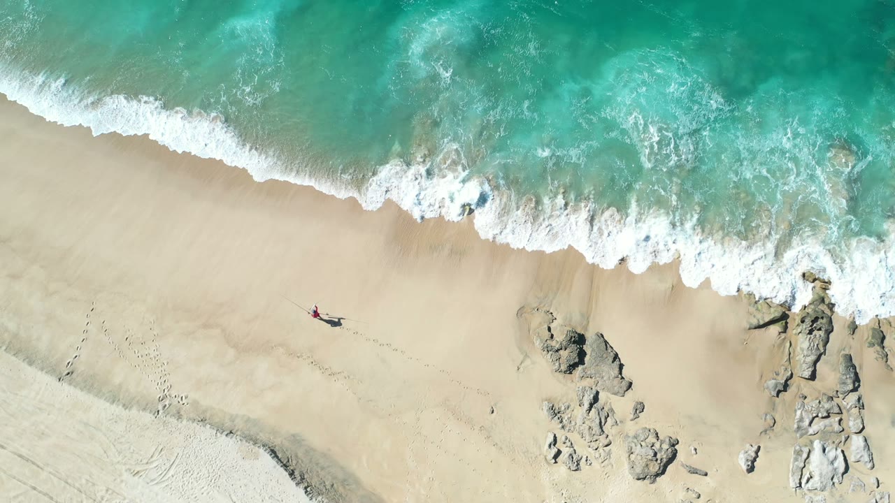 A high overhead Los Cabos beach frame shows one person on open sand, a diagonal band of white surf, and clusters of rocks near the edge of the water.