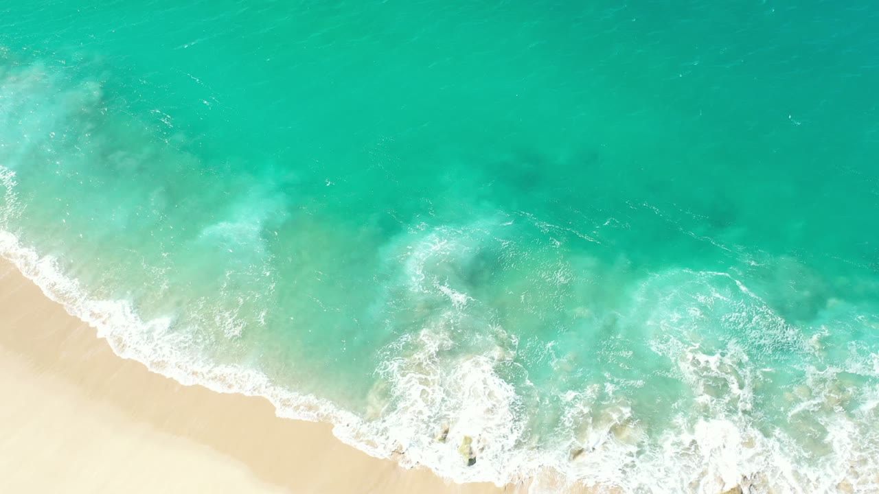A wider patch of foamy turquoise water churns near the lower sandy edge in Los Cabos, Mexico, with no visible people or structures.