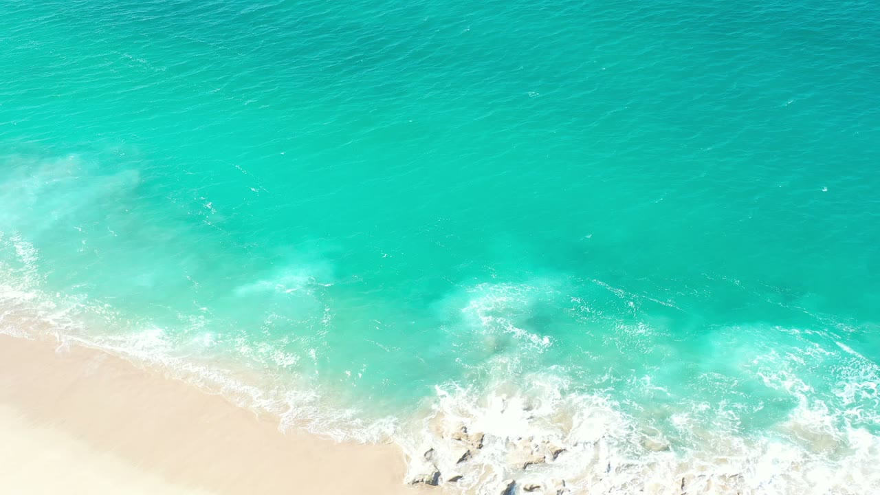 Aerial coastal frame in Los Cabos, Mexico, showing blue-green water, foamy wash, pale beach, and no visible people.