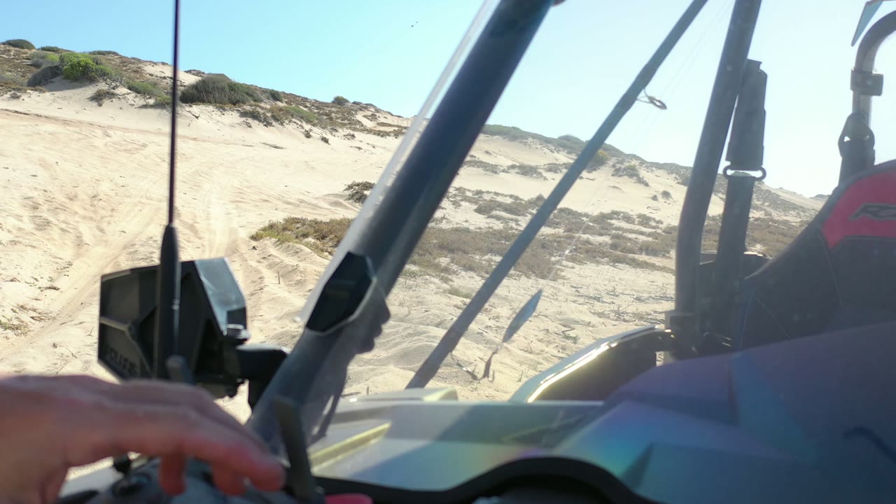 Interior POV in Los Cabos, Mexico, looking past vehicle bars and a black mounted device toward sunlit sand dunes, with a partial hand at the lower left.