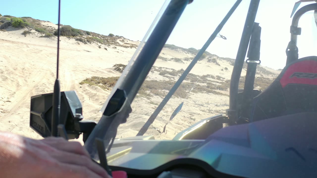 Los Cabos, Mexico view from inside or beside a small open vehicle, with diagonal bars and a black mount framing a sandy dune track and no visible face.