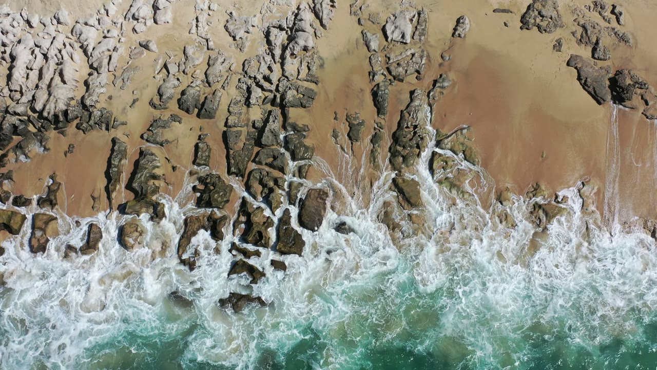 Overhead view in Los Cabos, Mexico, with white surf spreading over dark shoreline rocks and tan beach, no visible people.