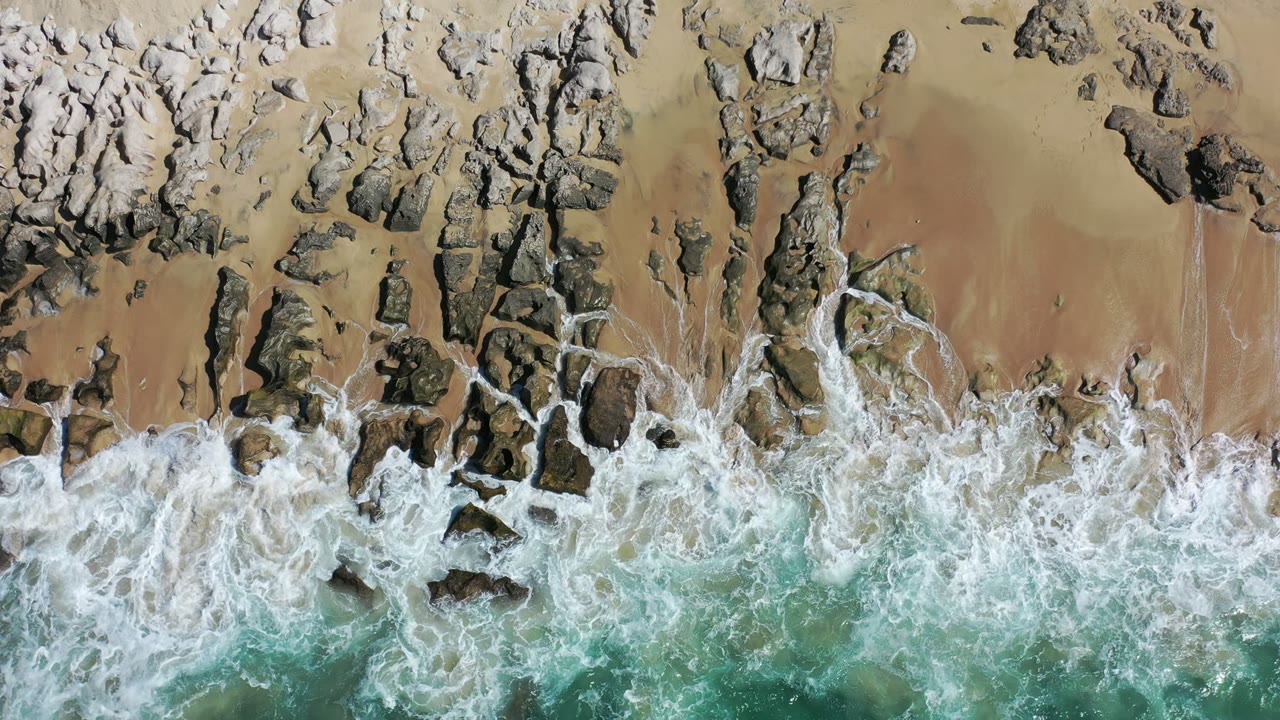 Aerial frame in Los Cabos, Mexico, showing a dense band of white foam curling around scattered dark rocks on tan sand, no visible people.