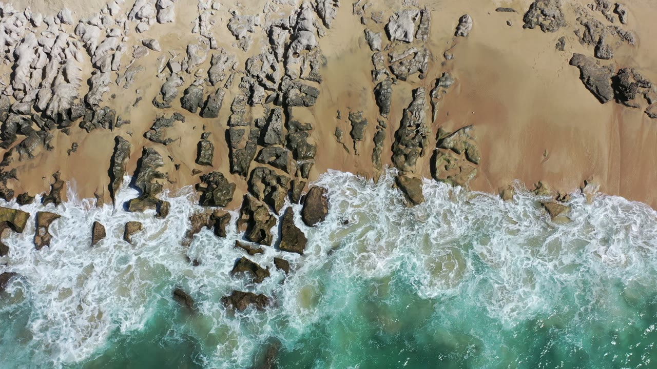 Aerial Los Cabos beach frame with white water pushed farther across the center rocks, tan sand above, turquoise sea below, and no visible people.