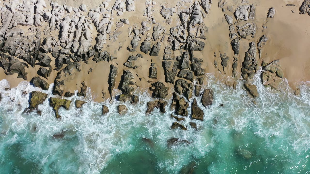 Top-down view of Los Cabos shoreline where white foam meets jagged rocks and tan sand, with turquoise water below and no visible people.