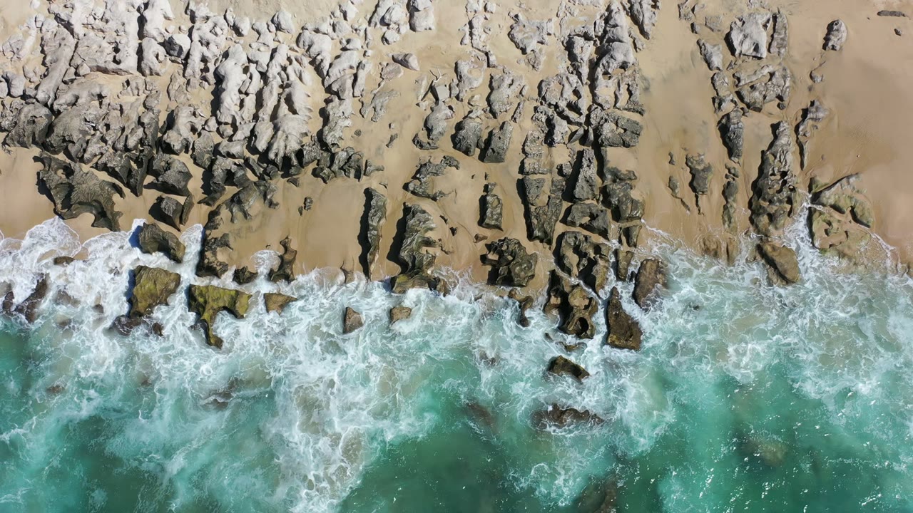Aerial Los Cabos beach frame with blue-green water pushing white foam around scattered dark rocks below a strip of sand, with no visible people.