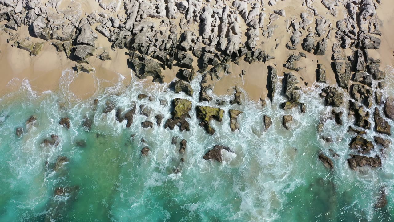 Foam gathers around scattered dark rocks as green water reaches the sandy Los Cabos shore, with no visible people.