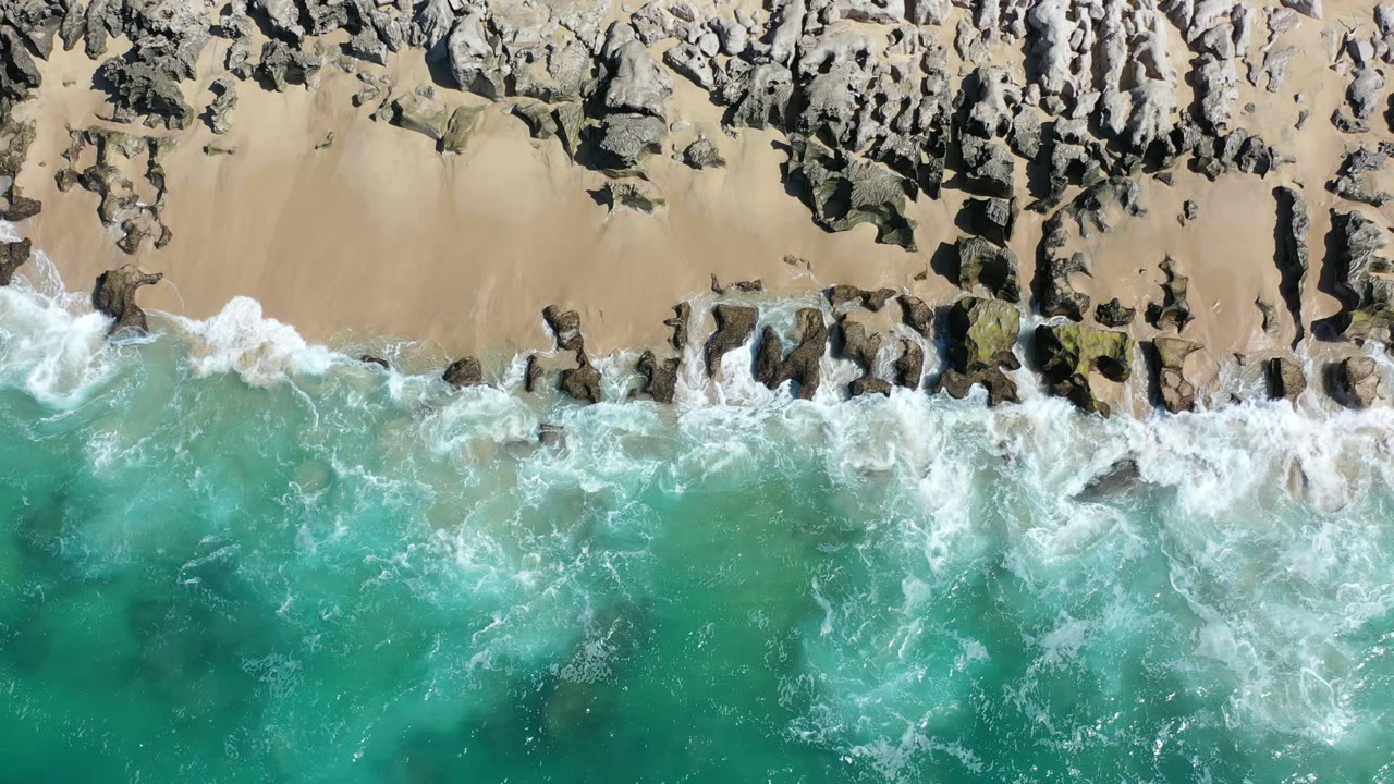 Overhead view in Los Cabos, Mexico, with turquoise water pushing white foam toward a rocky tan shoreline and no visible people.