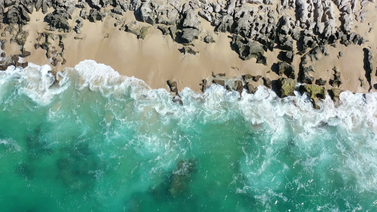 Overhead Los Cabos shoreline frame with a thicker band of white surf crossing shallow turquoise water below dark rocks, with no visible people.