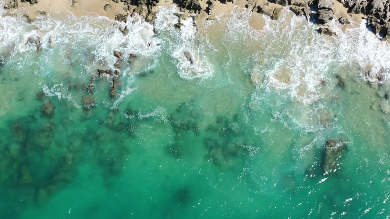 Overhead Los Cabos shoreline frame with foamy waves breaking across dark rocks near the sand and translucent green-blue water below, no visible people.