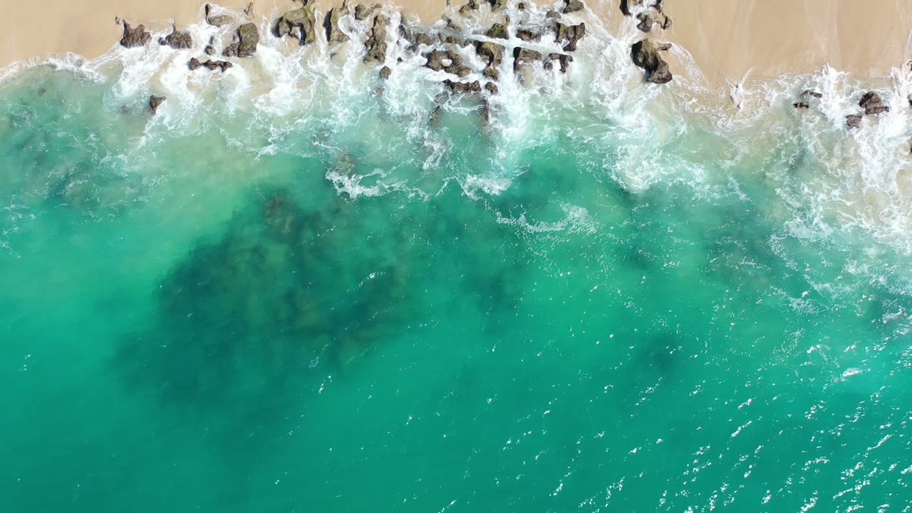 Aerial frame in Los Cabos, Mexico, showing a wider band of white foam crossing dark rocks above green-blue water, with no visible people.