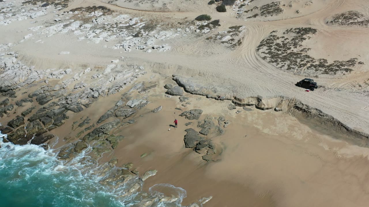 Aerial view in Los Cabos, Mexico, with blue-green surf entering from the lower left, dark rocks along the shore, tan sand, a black vehicle near the upper beach, and a small person in red near the rocks.