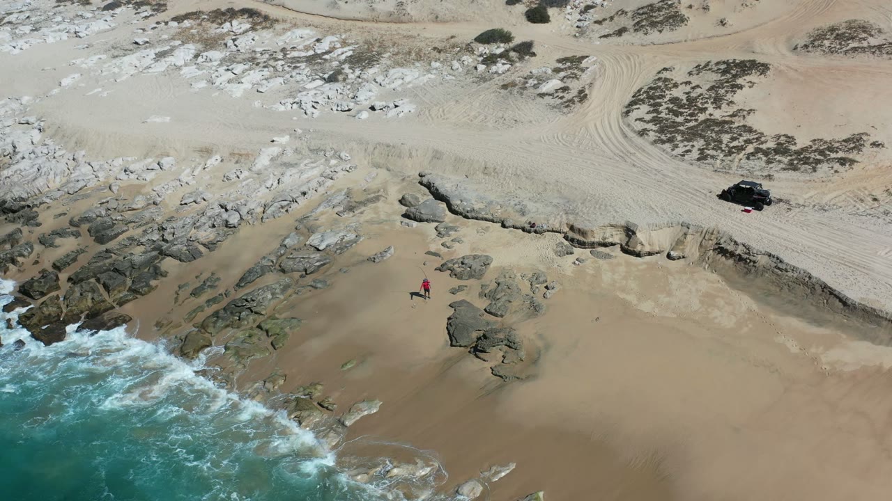 Aerial Los Cabos beach frame with more white water pushing against the left-side rocks, the red-clad person standing lower center-right on the sand, and the black vehicle still visible above the eroded beach edge.