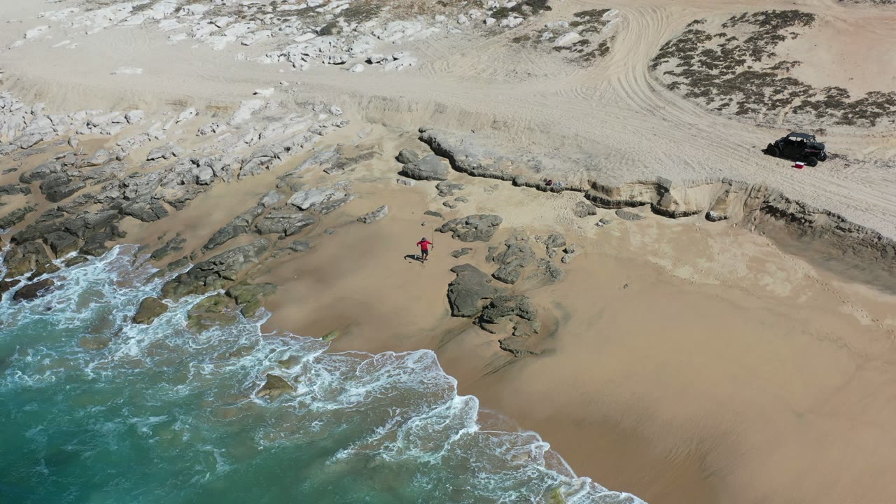 Aerial view of waves, rocks, sand, a small person, and a dark vehicle above the beach.