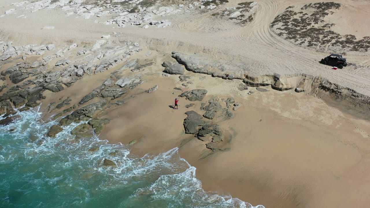 Aerial view in Los Cabos, Mexico, with foamy waves curling around exposed rocks as one person in red stands farther up the sandy beach below dune tracks.