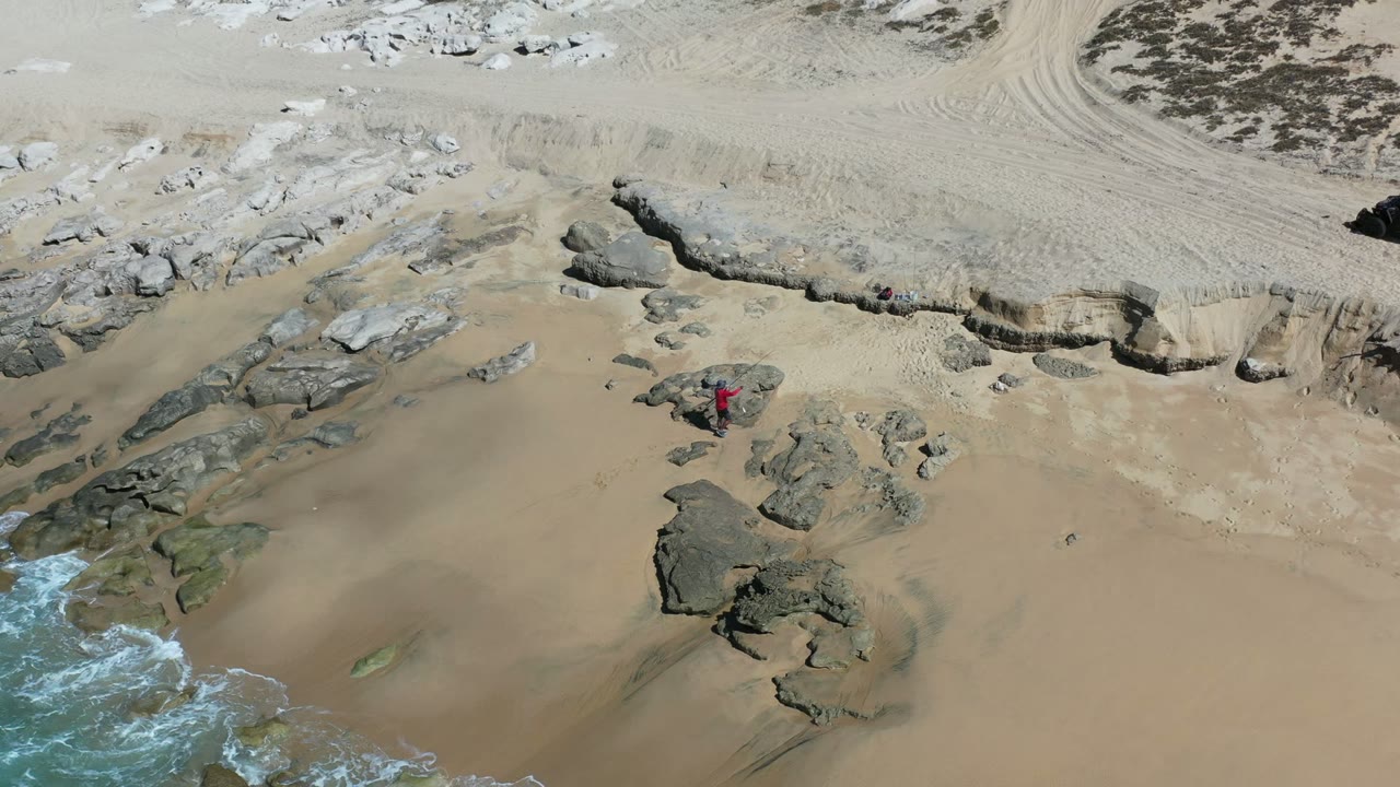 At the start of the Los Cabos clip, a fisherman in a red top stands near dark rocks on tan sand while a small edge of surf enters the lower left of frame.