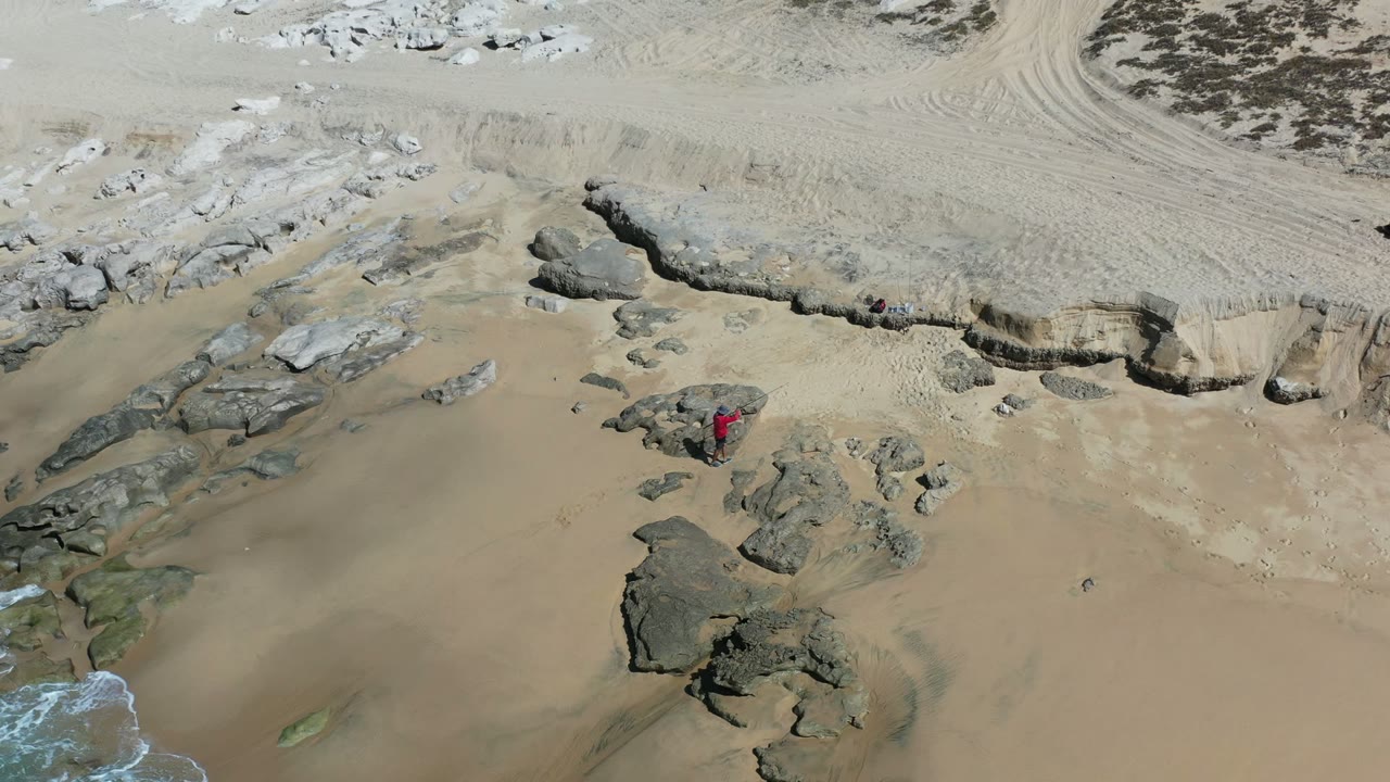 One second in, the Los Cabos fisherman appears slightly farther across the rocky sand, with pale water visible at the lower left and a carved sand bank across the upper frame.