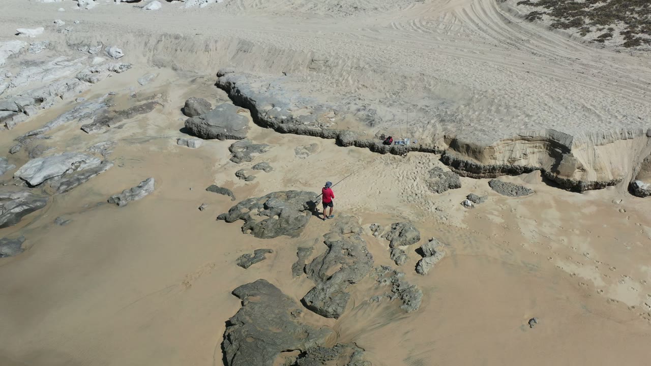 High aerial view in Los Cabos, Mexico, showing a child in a red shirt near scattered gray rocks on pale sand at the start of the clip.