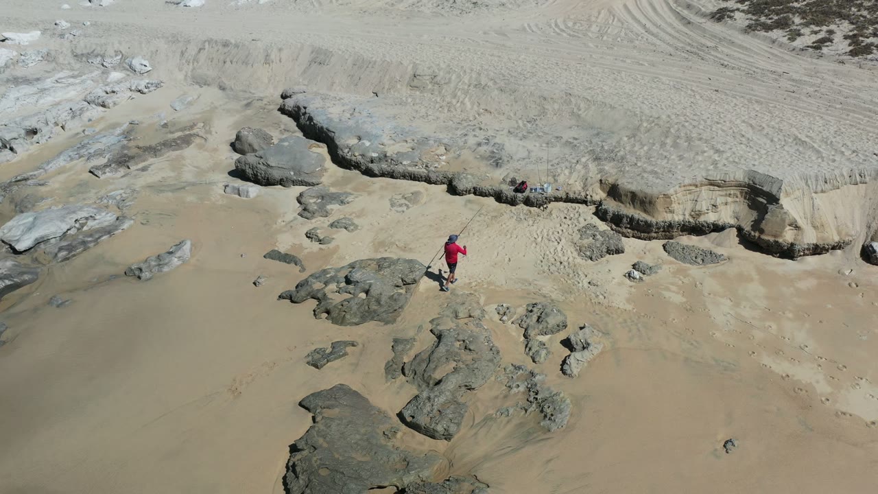 Aerial Los Cabos beach frame with the child slightly farther across the sand, walking beside flat gray rock shelves below an eroded sandy bank.
